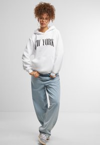 Felpa bianca con la scritta "NEW YORK" in nero, abbinata a jeans baggy di un blu chiaro. Il modello indossa occhiali rotondi e orecchini a cerchio in argento.