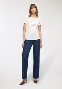Biała koszulka z okrągłym dekoltem i graficznym nadrukiem. Połączona z ciemnoniebieskimi szerokimi dżinsami i brązowymi klapkami. Gładka tekstura, luźny krój.