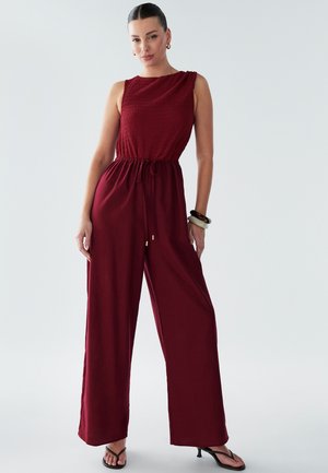 Burgundy jumpsuit with a textured, smocked bodice, wide-leg cut, cinched waist, and adjustable tie detail. Smooth fabric and minimal hardware.