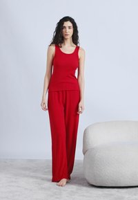 Γυναίκα με μακριά, σγουρά μαλλιά που φοράει κόκκινο αμάνικο τοπ και φαρδιά παντελόνια, όρθια ξυπόλητη σε χαλί δίπλα σε στρογγυλό λευκό καρέκλα.