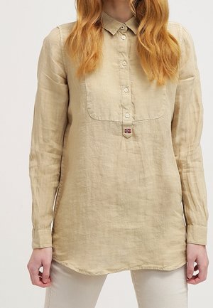 Femme portant une chemise en lin beige à manches longues boutonnée avec un petit patch représentant un drapeau près du bouton inférieur.