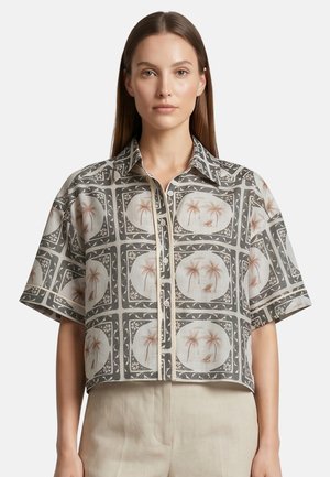 Donna con capelli castani lisci indossa una camicia ampia a maniche corte con bottoni e stampa di palme e uccelli, abbinata a pantaloni beige chiaro.