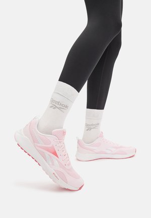 Jasnoróżowe buty sportowe z panelami z siateczki i białymi detalami, z amortyzowaną podeszwą, podkreślone różową gumową podeszwą. Noszone z czarnymi legginsami i białymi skarpetkami.