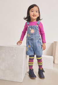 Peto vaquero con un parche floral, combinado con una camisa de manga larga rosa. Medias a rayas multicolores y botas azul marino con detalles de borlas.