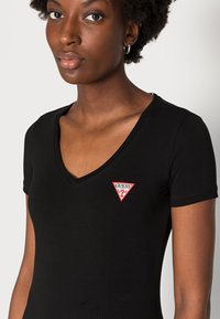 T-shirt nera attillata con scollo a V in tessuto morbido, caratterizzata da un piccolo logo triangolare rosso e bianco sul lato sinistro del petto.