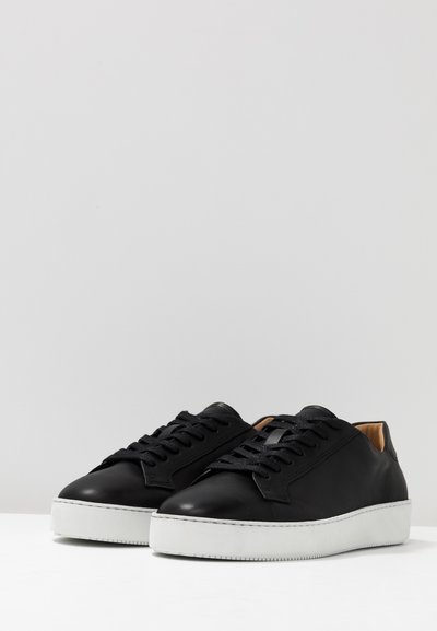 Sneakers in pelle nera con suola in gomma bianca piatta, punta rotonda, chiusura con lacci davanti e texture liscia. Interno in pelle marrone chiaro. Design minimalista.