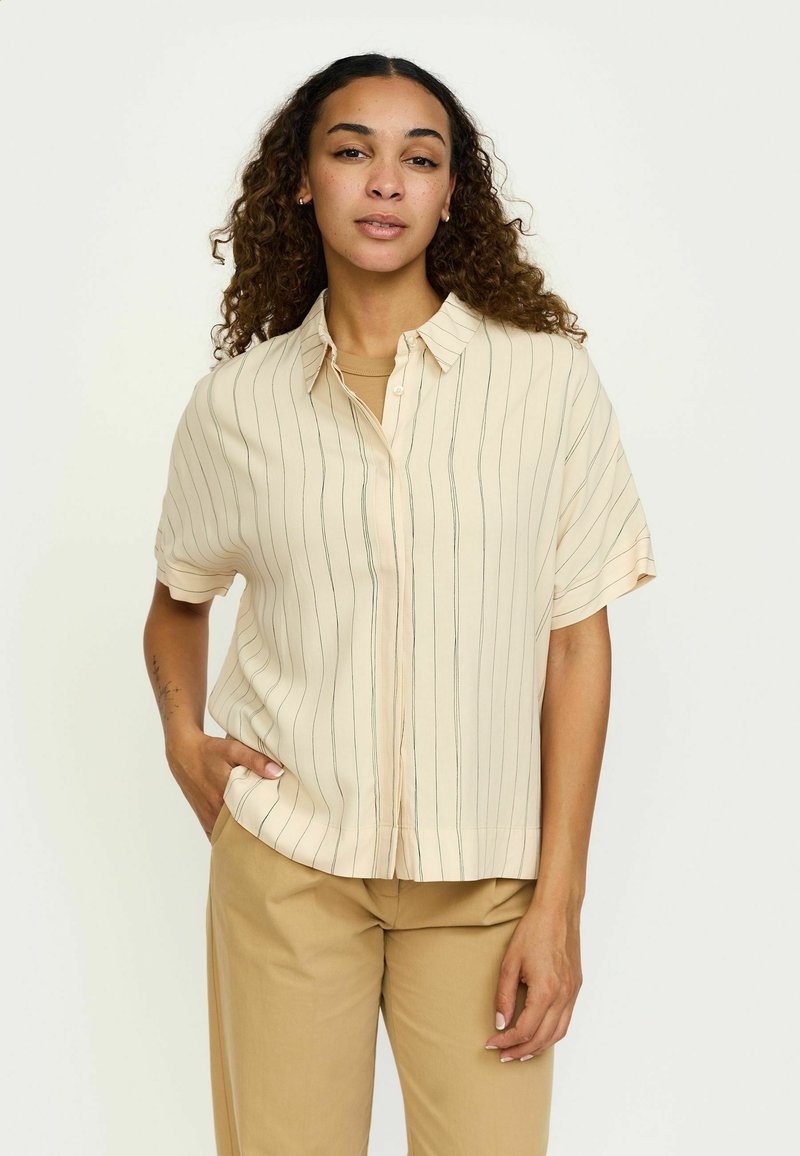 Short-sleeve, cream blouse with vertical green stripes, button-down front, and relaxed fit. Styled with beige trousers.