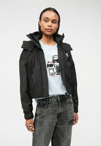 Juodas trumpas švarkas su aukštu apykakle ir užtrauktukų kišenėmis, dėvimas virš šviesiai mėlynos spalvos marškinėlių su animaciniu piešiniu. Textūruoti pilki džinsai.