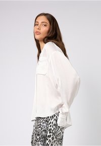 Hvit bluse med lange ermer og en frontlomme; glatt tekstur. Kombinert med sølvfargede leopardmønstrede paljettbukser med innsvingt midje.