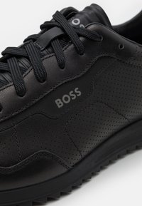 Chaussures de sport noires en cuir lisse avec des parties perforées. Design à lacets avec semelle en caoutchouc et logo visible sur le côté.