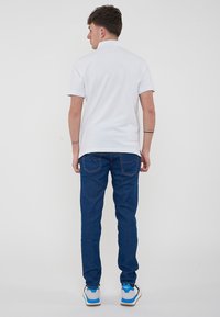 Polo shirt bianco a maniche corte, jeans blu scuro con tasche posteriori, e sneakers con dettagli blu, mostrato da dietro.