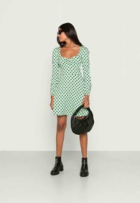 Vestido verde menta com bolinhas, mangas longas e decote quadrado. Uma bolsa de mão preta acolchoada e botas de tornozelo pretas, robustas, complementam o look.