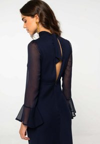 Vestido azul marino con mangas largas transparentes, acentos con volantes y diseño de corte en la espalda. Confeccionado con tela ligera de acabado texturizado.