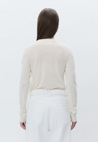 Top beige clair à manches longues en tissu doux, silhouette ajustée, avec un col rond, associé à un jean blanc taille haute, poches arrière visibles.