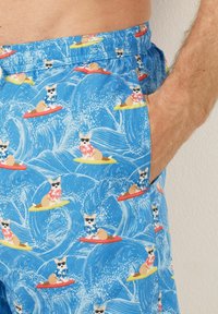 Shorts de bain bleus ornés d'un design de chien de dessin animé, surfant sur des vagues, avec des chemises à fleurs en rouge et bleu. Taille élastique avec poches latérales.