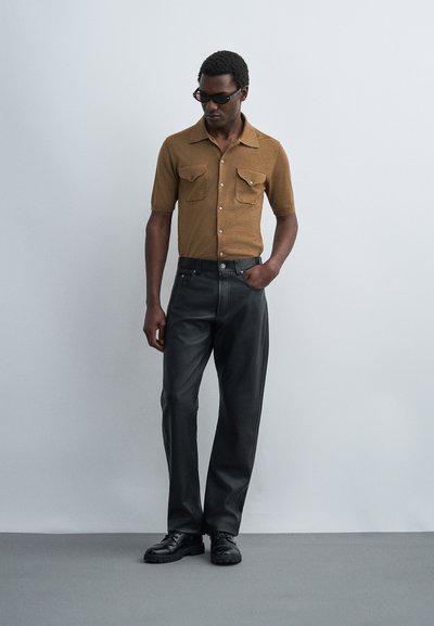 Άνδρας που φοράει μαύρα γυαλιά ηλίου, καφέ κοντομάνικο πουκάμισο με κουμπιά, μαύρο δερμάτινο παντελόνι και μαύρα παπούτσια, στέκεται με το ένα χέρι στην τσέπη.