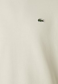 Ljusbeige bomull T-shirt med rund halsringning och en liten grön krokodil-logotyp på vänster bröst. Slät textur, enkel design.