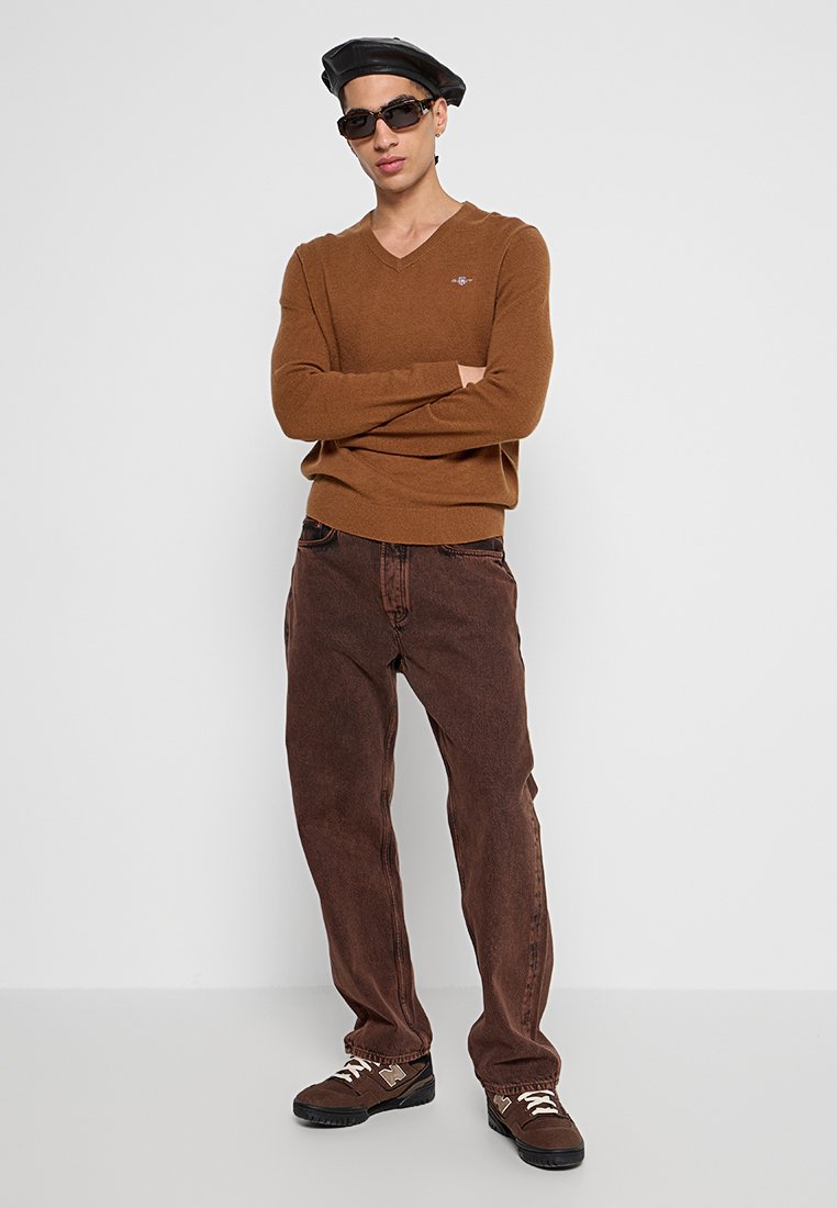 Brauner V-Ausschnitt-Pullover mit strukturiertem Finish, kombiniert mit dunkelbraunen, locker sitzenden Jeans. Akzente sind ein kleines Logo auf dem Pullover und schwarze Sonnenbrillen.
