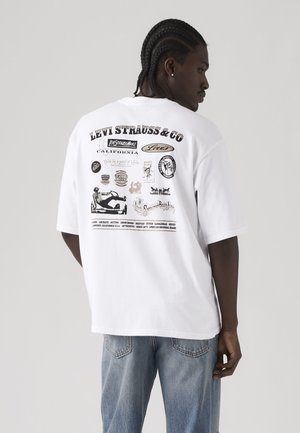 Hombre con trenza, lleva una camiseta blanca de Levi's con logotipos y texto vintage en la parte trasera, combinada con vaqueros azul claro, de espaldas.