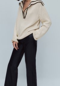 Pull en maille beige avec un large col, des finitions noires et des détails de boutons ; associé à un pantalon noir, brillant et à jambes larges.