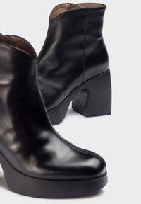 Botas de tobillo de cuero negro con un acabado brillante, que cuentan con un tacón robusto negro y contornos suaves. Cremallera lateral para un fácil uso.