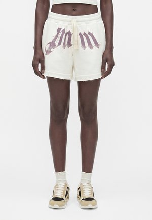 Γυναικείο μοντέλο που φοράει λευκά σορτς με κορδόνι και μεγάλα γοτθικού στυλ γράμματα, συνδυασμένα με λευκές κάλτσες και μπεζ-μαύρα αθλητικά παπούτσια.