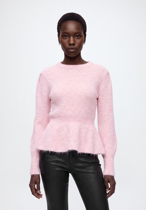Femme portant un pull texturé rose clair à manches longues avec un ourlet péplum et un pantalon en cuir noir, debout devant un fond blanc uni.