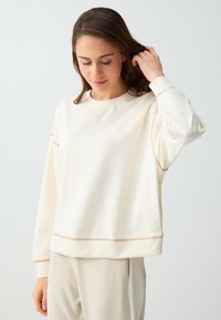 Jeune femme portant un sweat crème et un pantalon assorti, touchant ses cheveux et regardant vers le bas sur un fond clair uni.