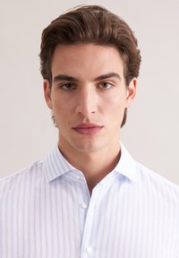 Koszula na guziki w jasnoniebiesko-białe paski z kołnierzem rozłożystym. Tkanina jest gładka z subtelnym połyskiem i posiada małe białe guziki.