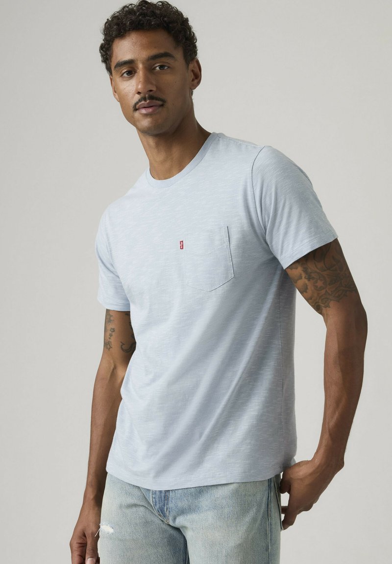 Uomo con baffi che indossa una t-shirt a maniche corte azzurro chiaro con tasca e jeans azzurri chiari, che mostra tatuaggi su entrambe le braccia su sfondo neutro.