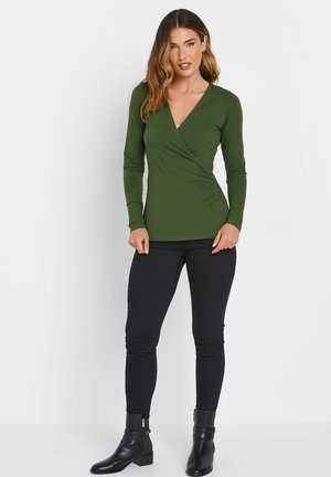 Haut à manches longues et col en V en tissu vert foncé avec un design croisé et un ruché sur le côté, associé à un pantalon skinny noir et des bottines.