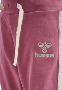 Lila sweatpants med en strukturerad yta, med en krämfärgad dragsko och en silverbroderad logo på låret. Sidosträck har ett geometriskt mönster.