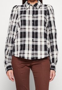 Chemise à carreaux boutonnée avec un motif à carreaux noir, blanc et marron. Caractérisée par des manches bouffantes et un col classique. Tissu doux et texturé.