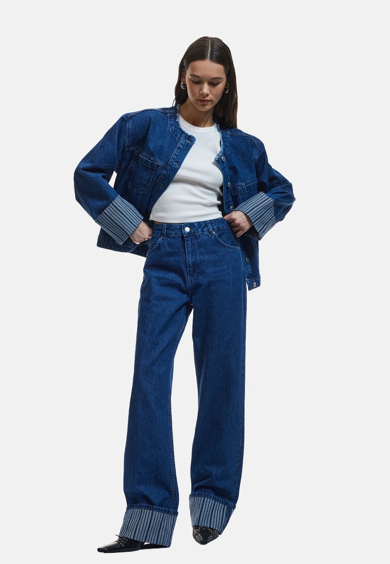 Chaqueta de denim y jeans de tiro alto en azul oscuro con puños a rayas. El modelo lleva una blusa blanca. Los zapatos presentan un diseño texturizado en negro.