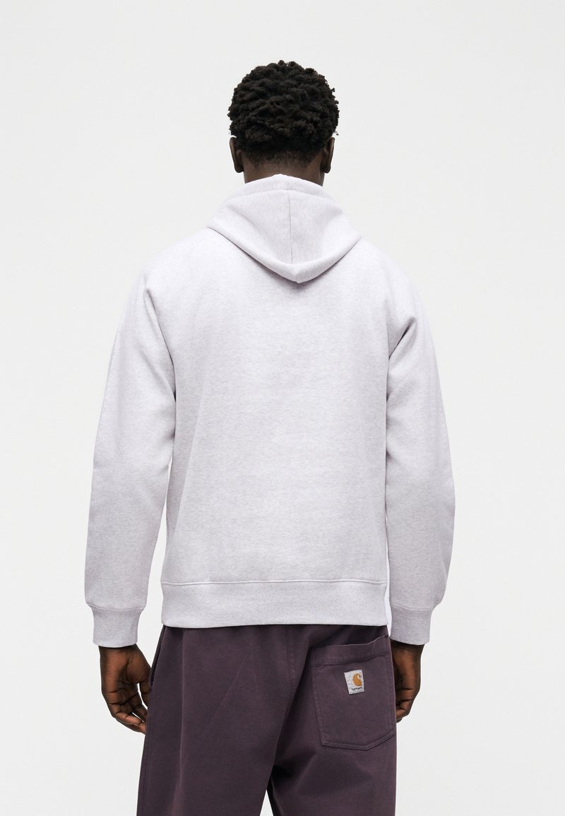 Man met donker krullend haar, gekleed in een lichtgrijze hoodie en donkerpaarse broek, gezien van achteren tegen een effen witte achtergrond.