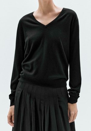 Personne portant un pull noir à col en V manches longues et une jupe plissée noire sur un fond clair uni.
