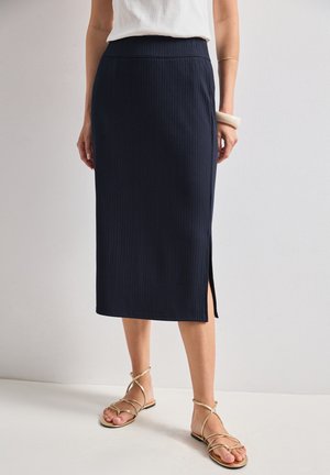 Femme portant une jupe midi côtelée bleu marine avec une fente latérale, un haut blanc, des bracelets dorés et des sandales plates dorées à brides, debout sur un sol blanc.
