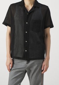 Maglietta nera a maniche corte realizzata in tessuto a rete testurizzato, con taschino e chiusura a bottoni. Indossata con jeans grigi, presenta una vestibilità comoda.