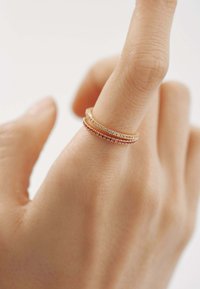Anillos de oro apilados en un dedo. Un anillo cuenta con piedras preciosas transparentes, mientras que otro tiene acentos rojos. La mano tiene un tono de piel claro y está posicionada contra un fondo blanco.