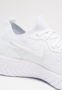 Chaussure de course blanche avec un upper en tricot et une semelle texturée. Présente un swoosh blanc proéminent et un système de laçage. Design lisse et sans couture.