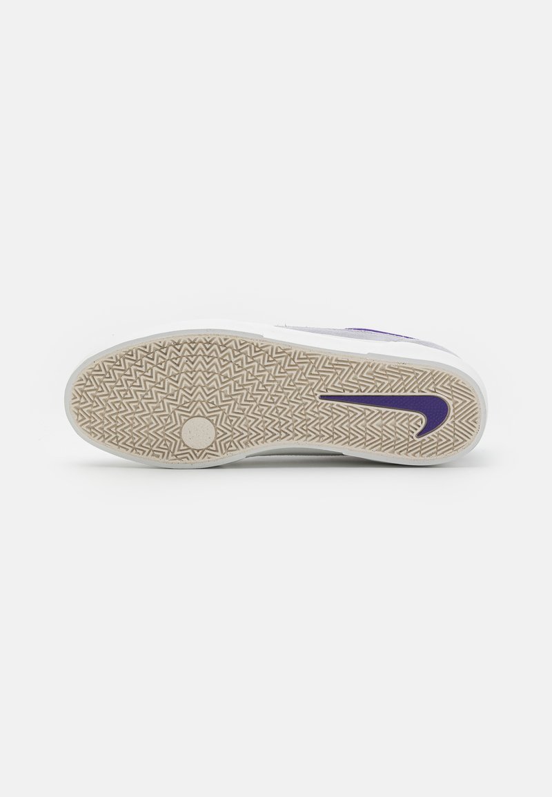 Weißer Sneaker mit einer strukturierten Gummisohle, die ein Zickzackmuster aufweist und ein lila Nike-Swoosh zeigt. Glatter oberer Bereich sichtbar.