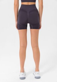 Hochtaillierte, enganliegende Shorts aus dunkelviolettem Stoff mit strukturierten Einsätzen und kontrastierenden Nähten. Kombiniert mit weißen Sportschuhen.