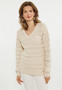 Beige gebreide trui met een V-hals, voorzien van een gestructureerd, patroonontwerp met diamantvormen en ribbels aan de mouwen. Gemaakt van lichtgewicht materiaal.