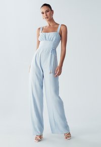 Svetlo modra kombinezona z kvadratnim izrezom, nabranim bustom, pasom z obročem in širokimi hlačami. Narejena iz gladkega materiala z subtilnim sijajem.