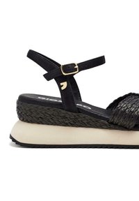 Sandalias de plataforma trenzadas en negro con una suela gruesa beige, correa ajustable con herrajes dorados y tejido texturizado en la parte superior.