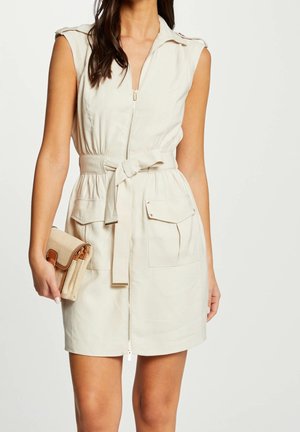 Beżowa sukienka bez rękawów z przednim zamkiem na suwak, wiązanym paskiem w talii, kieszeniami z klapkami oraz dopasowaną beżową kopertówką trzymaną w lewej ręce.