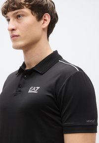 Svart polotrøye med ribbet krage, med hvit EA7-logo og sidestriper, laget av teksturert stoff. Korte ermer med "VENTUS"-detalj.