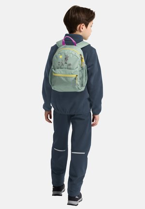 Jongen gekleed in een marineblauw tenue en een lichtgroene rugzak met diermotieven, staand en met de rug naar voren tegen een effen achtergrond.