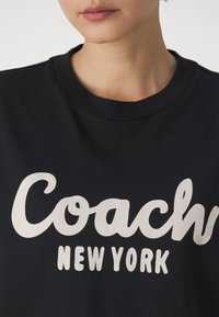 Zwarte katoenen t-shirt met grote witte tekst: "Coach" en "NEW YORK." Ronde hals, gladde textuur, close-up van het borstgebied.