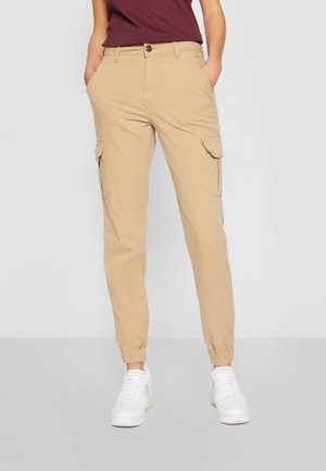 Pantalones cargo beige con puños elásticos usados con camiseta burdeos y zapatillas blancas, manos en los bolsillos, sobre un fondo claro y liso.